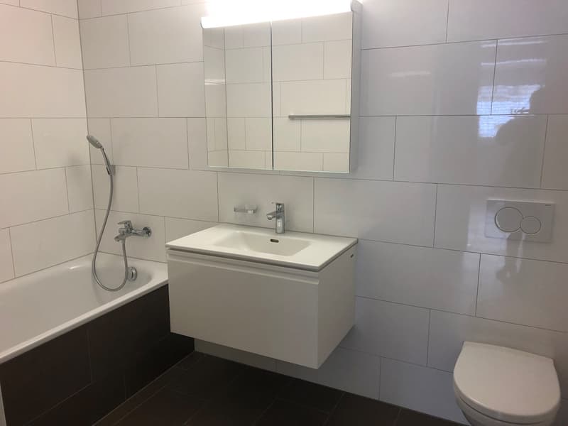 The image shows a modern bathroom with white tiles on the walls and floor, a vanity unit with a sink, a mirror cabinet, and a toilet. The bathroom appears to be well-lit and has a clean, minimalist design.