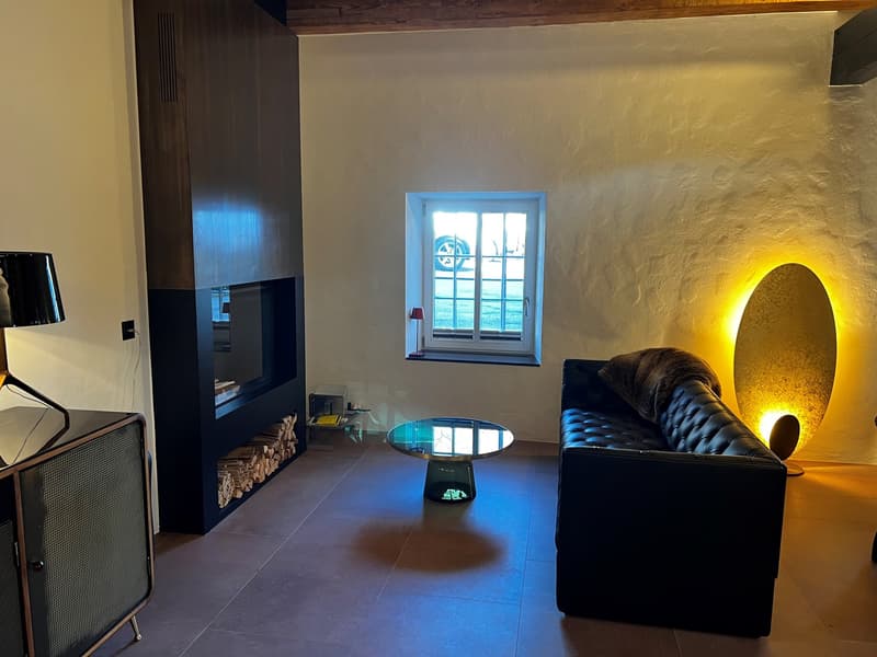 Cozy living room with a fireplace, black leather sofa, glass coffee table, and wooden accents. The room has a warm, inviting atmosphere with a large window providing natural light and a decorative lighting fixture on the wall.