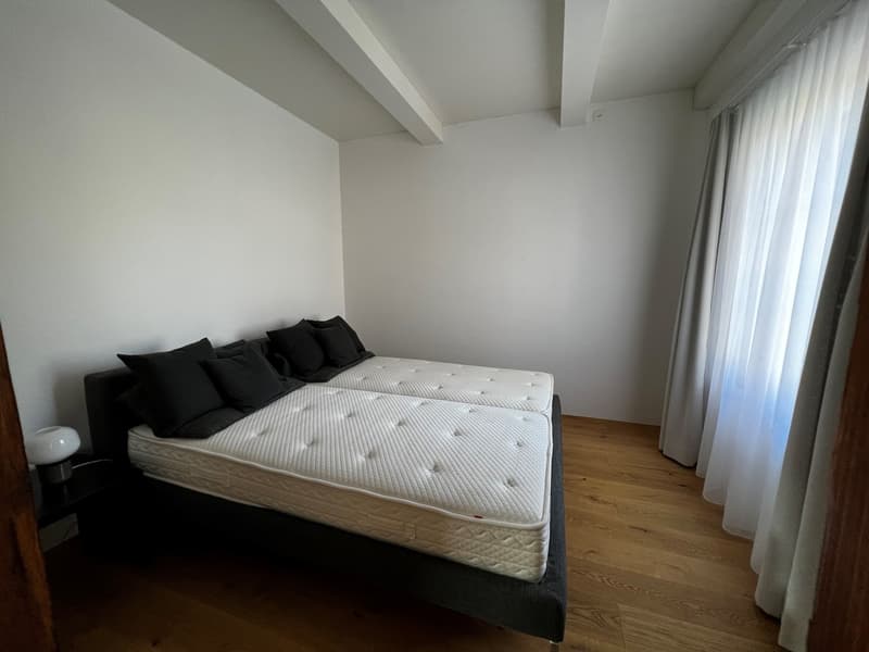 Spacious bedroom with white walls, hardwood flooring, a large bed with a white mattress and black bedding, and curtains covering the windows.