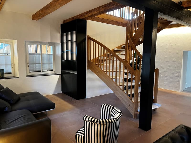 Open-concept living space with wooden beams, black and white striped armchair, black leather sofa, wooden staircase, and built-in shelving unit