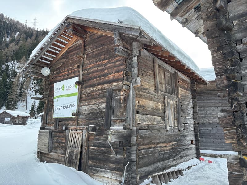 Rustic wooden cabin in a snowy mountain setting, with a 'For Sale' sign displayed on the exterior.