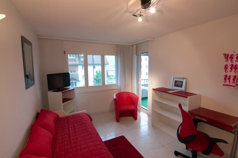 A small furnished room with a red sofa bed, red chair, and a desk with a red chair. The room has white walls and tiled flooring. There is a television on a shelf, a mirror, and a picture frame on the wall.