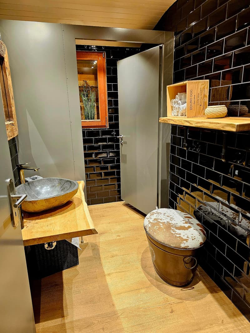 The image shows a rustic-style bathroom with black subway tile walls, a wooden vanity with a round stone sink, and a wooden shelf with decorative items. The floor appears to be made of wood planks. The bathroom has a unique and cozy atmosphere.