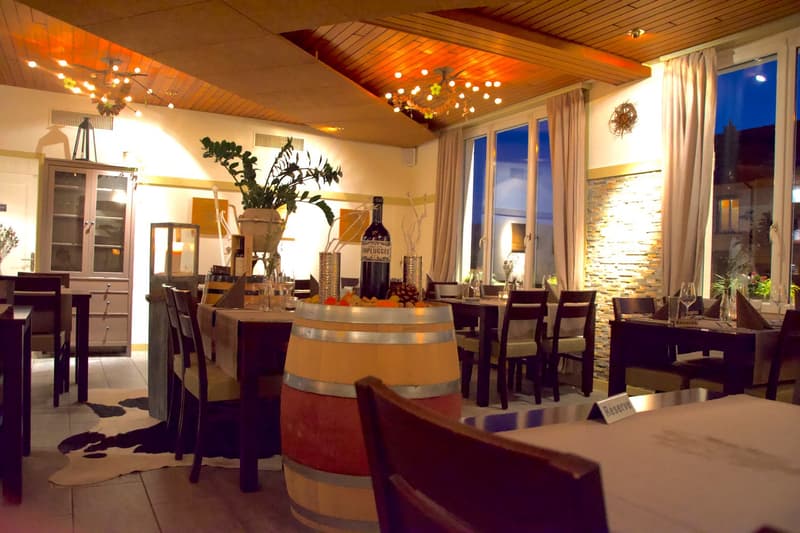 Spacious dining room with wooden ceiling, large windows, chandelier lighting, wooden dining table and chairs, wine barrel decor, and a fireplace visible.