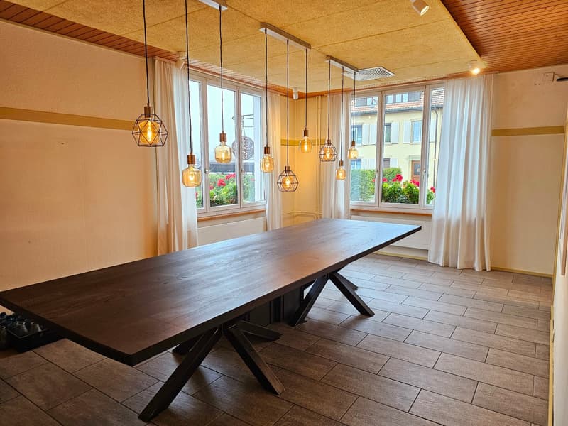 Large wooden dining table with a modern, geometric light fixture above it. The room has large windows overlooking a garden or outdoor area. The walls are a warm, neutral color and the floor is tiled.