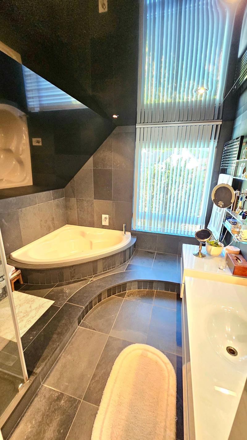 Spacious bathroom with a corner bathtub, tiled floors, and a large window providing natural light. The bathroom also features a vanity with a mirror and some decor items.