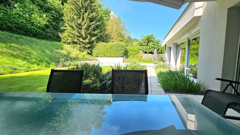 The image shows a modern outdoor living space with a swimming pool surrounded by lush greenery. There is a covered patio area with comfortable seating overlooking the pool. The property appears to be situated on a hillside, providing a scenic view of the