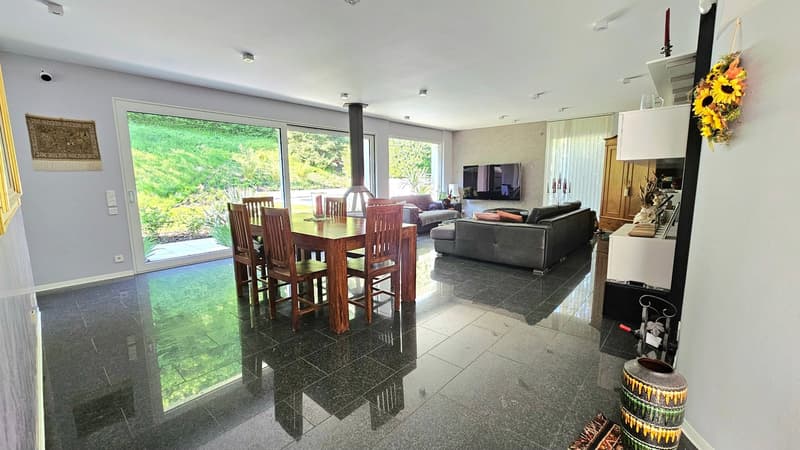 Spacious open-plan living and dining area with large windows overlooking a garden, hardwood dining table and chairs, grey sofa, fireplace, and decorative elements like a sunflower arrangement.