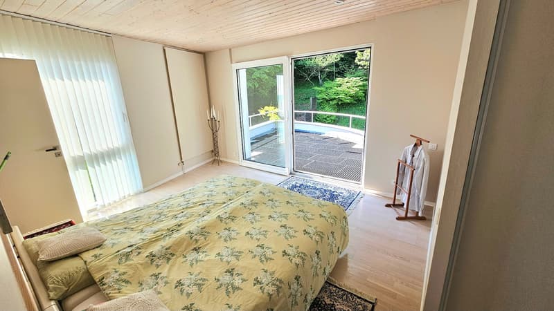 Spacious bedroom with large windows overlooking a balcony and lush greenery outside, hardwood floors, floral patterned bedding, and a standing lamp