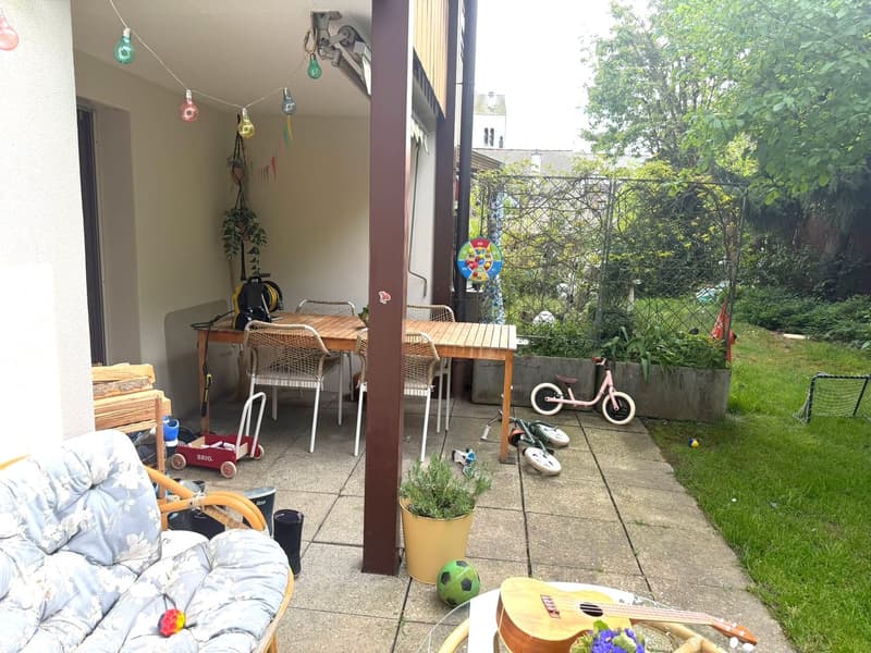 Outdoor area with wooden table, chairs, plants, and toys. A small pink bicycle is parked on the side.