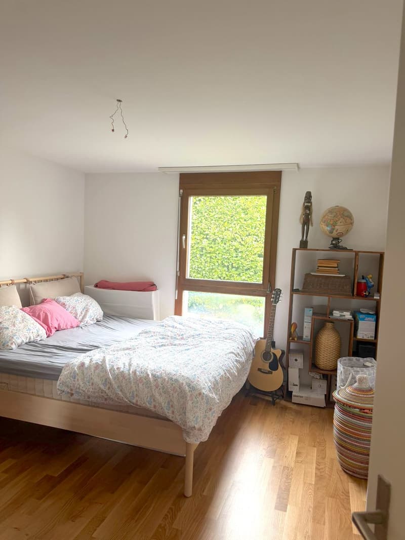 Bedroom with bed, wooden floor, white walls, large window, guitar, bookshelf, storage box, globe, decorative items