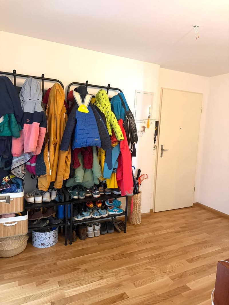 A room with a wooden floor, a white door, a white wall, a black metal rack with clothes and shoes, a basket, and a wicker basket.