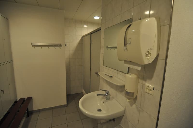 A small bathroom with white tiles, a white sink, a mirror, a hand dryer, a soap dispenser, and a bench.