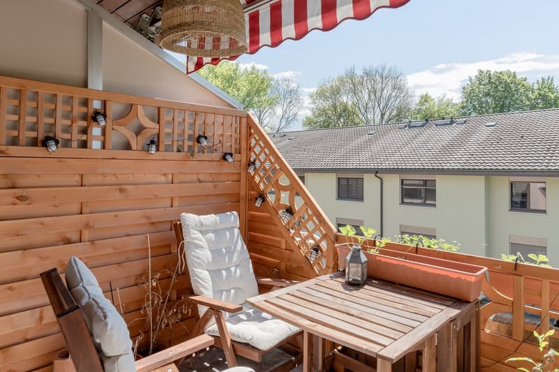 Wooden balcony with a striped awning, outdoor furniture including a table and chairs, potted plants, and string lights. The balcony overlooks a residential building with trees in the background.