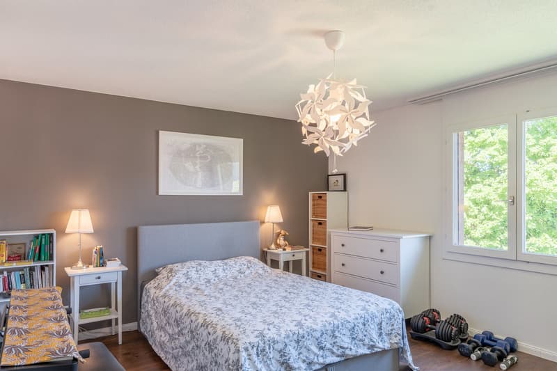 Bedroom with bed, two bedside tables with lamps, white bookshelf, wardrobe, two dumbbells on floor, chandelier, large window, framed photo on wall