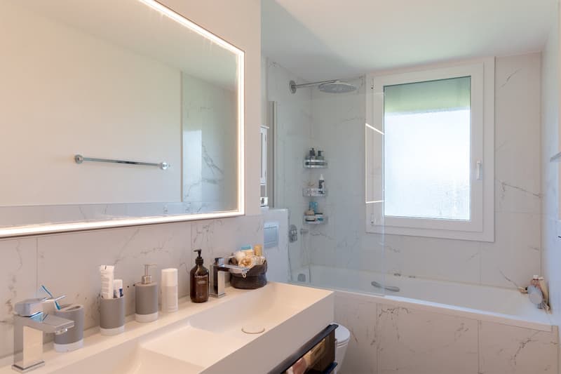 Spacious bathroom with marble-like tiles, large mirror, vanity with sink, shower area with glass enclosure, and shelving for toiletries