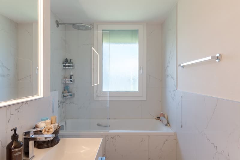Spacious bathroom with marble-like tiles, a bathtub, a shower, a window, and various bathroom accessories and shelves