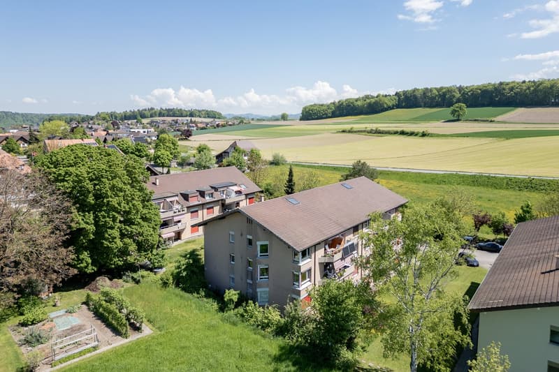 The image shows a scenic rural landscape with a small town or village nestled among rolling hills and fields. The buildings appear to be multi-story residential apartments or condominiums surrounded by lush greenery and trees. The landscape features a mix