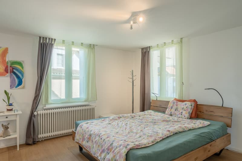 wooden bed, double bed, wooden bed frame, pillows, bedspread, small nightstand, potted plant, clock, white radiator, artwork, tall windows, curtains