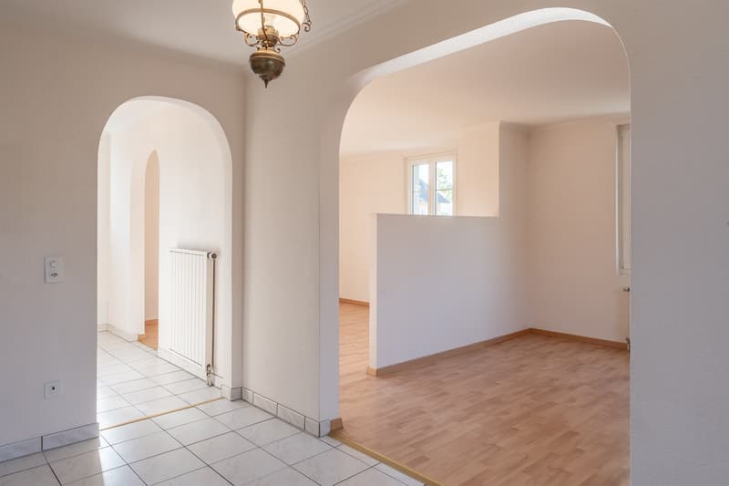 Empty room with white walls, wooden floor, arched doorways, white ceiling, white radiator, power outlets, and a window.