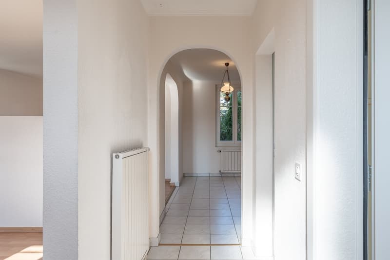 Empty hallway with white tiles, white walls, arched ceiling, radiators, and a window with a view outside.