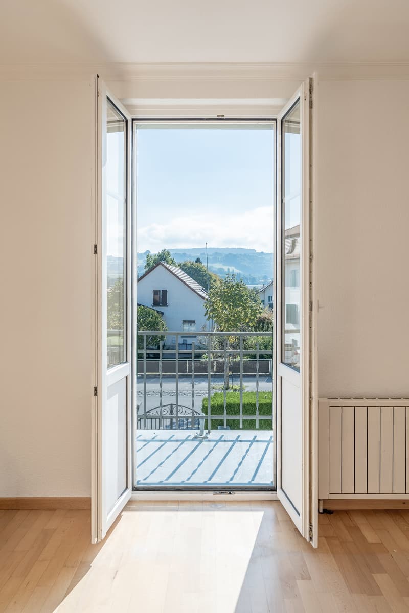 Open door leading to a balcony with a metal railing, wooden floor, white walls, and a view of a house and mountains in the distance