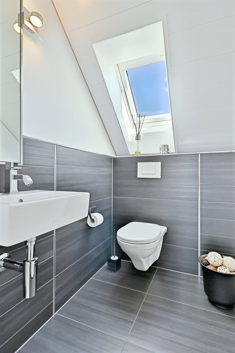A modern bathroom with a white sink, white toilet, gray walls, a window, and silver decorative balls in a black bucket.