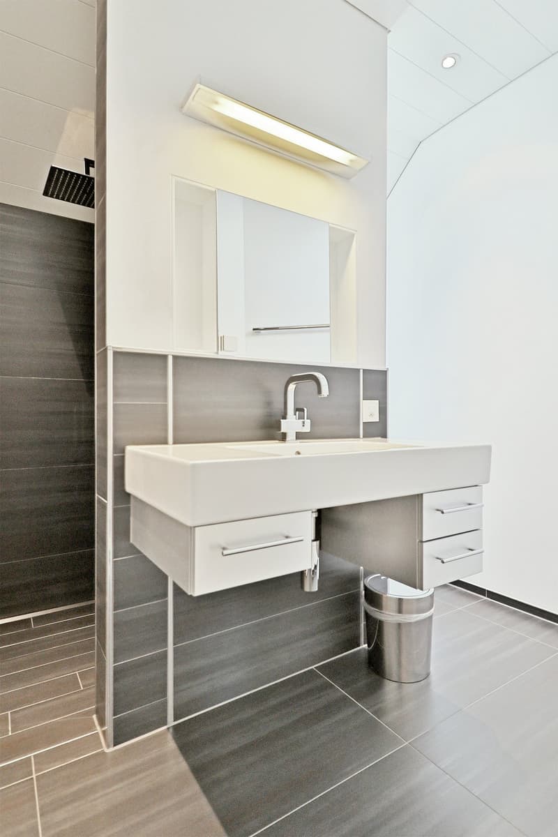 A modern bathroom with white tiled walls, a white countertop, stainless steel fixtures, and a shower stall.