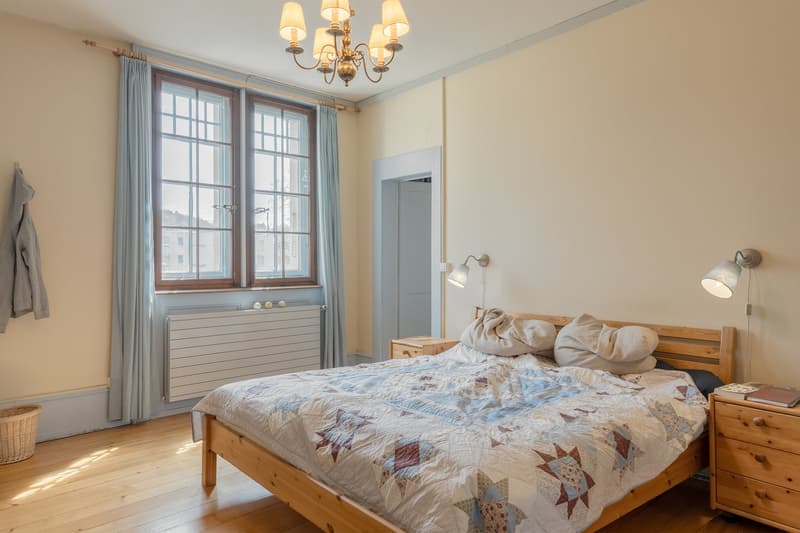 wooden bed, quilt, wooden nightstands, chandelier, radiator, window with curtains, wicker basket