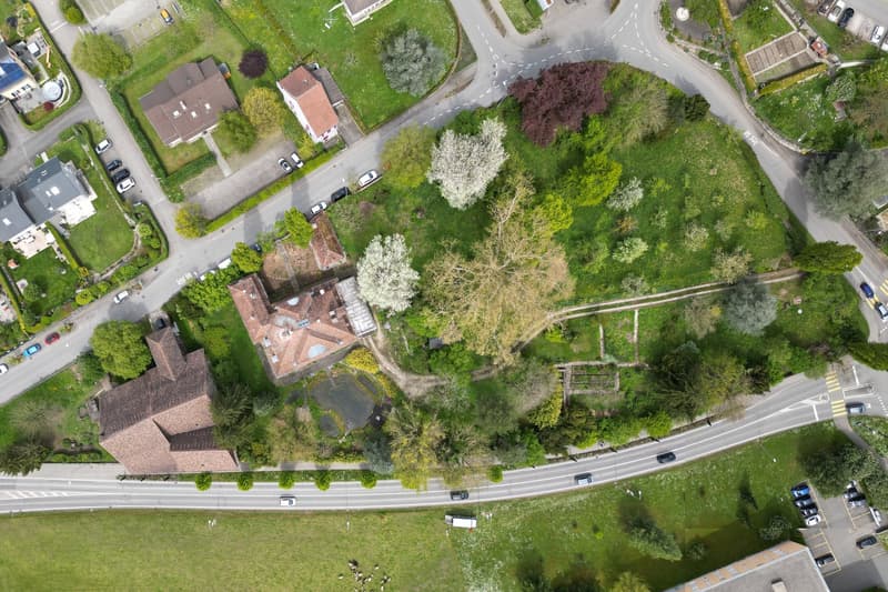 Aerial view of a neighborhood with several houses, roads, and greenery. The houses have different architectural styles and are surrounded by trees and grass. There are cars parked in the driveways and on the side of the road.