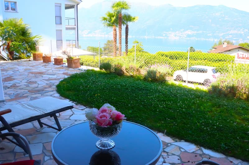 stone patio, lawn, 2 lounge chairs, table with flower arrangement, white car parked outside fence, fenced garden, mountains and lake in distance