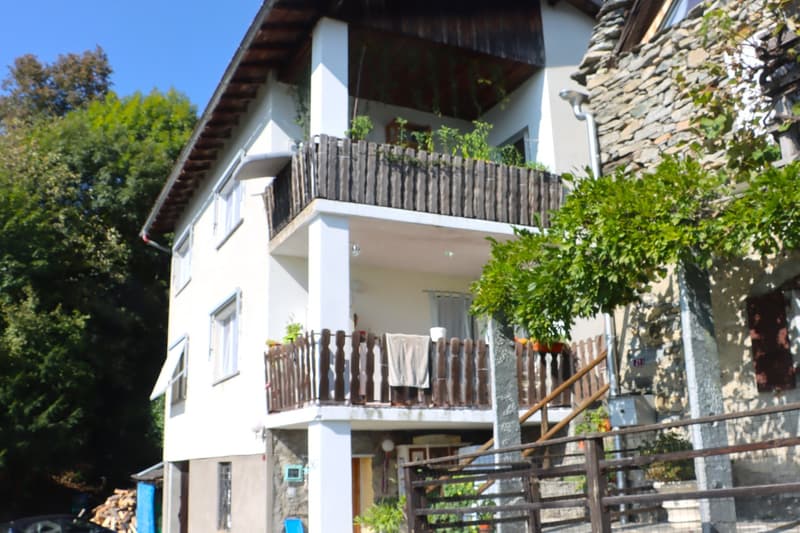 2 story house, white exterior, wooden balcony, stone wall, trees in the background