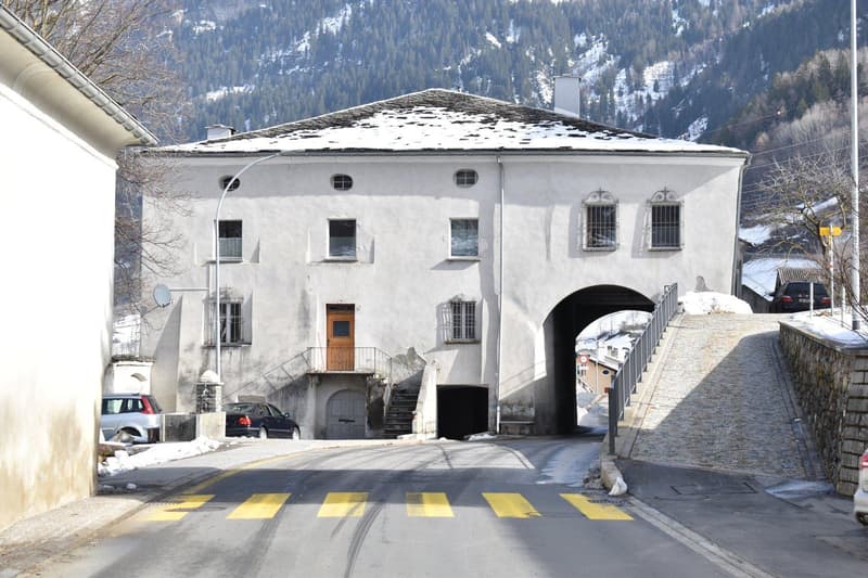 Large building with multiple windows, entrance with stairs, attached garage, snow covered roof, mountain in the background