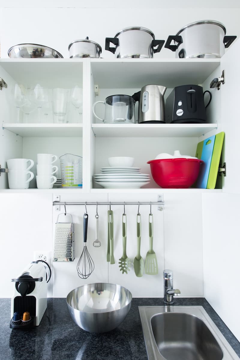 Well-organized kitchen with shelves displaying various kitchen appliances and utensils, including pots, pans, cups, and a coffee maker. The kitchen also features a sink, a stainless steel bowl, and a rack for hanging cooking tools.