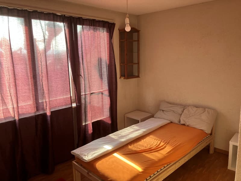 Single bed, wooden floor, white walls, window with curtains, small table, wooden cabinet