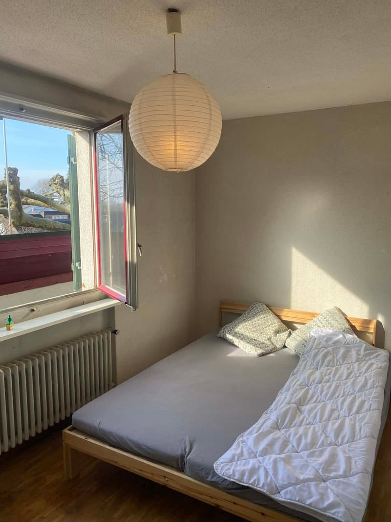 A bedroom with a bed, pillows, and a blanket. A radiator is on the wall. A window with a view of the outside. A lamp is hanging from the ceiling.