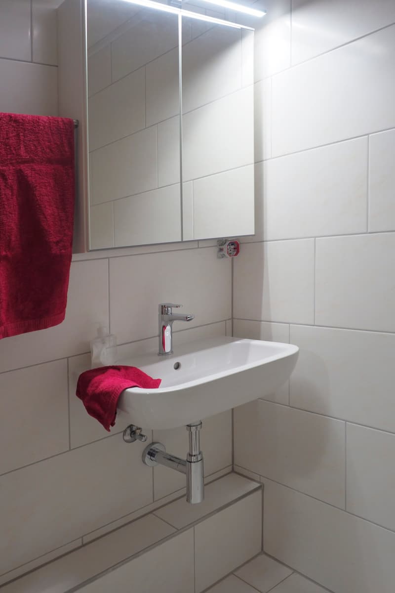 Modern, compact bathroom with white tiles, modern mirror, white sink, chrome faucet, towel, and tiled shower bench.
