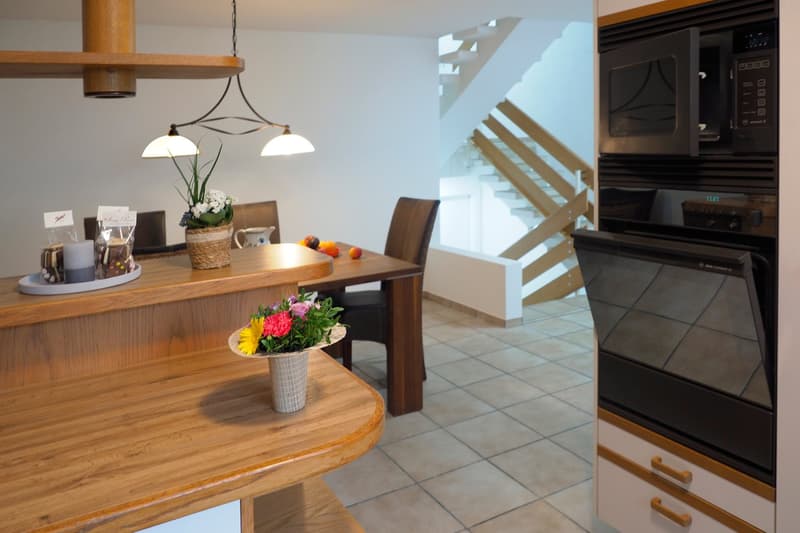 modern wooden kitchen with a breakfast bar, dining table, chairs, flowers, wooden staircase in the background, and kitchen appliances