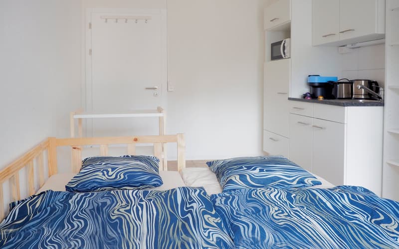 small bedroom with a double bed, white walls, wooden headboard, wooden baby crib, kitchen area with microwave and sink
