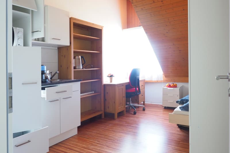 Wooden desk with black office chair, desk lamp, shelf, wooden floor, open kitchen with kettle, sink, microwave, bedroom with bed, small dresser