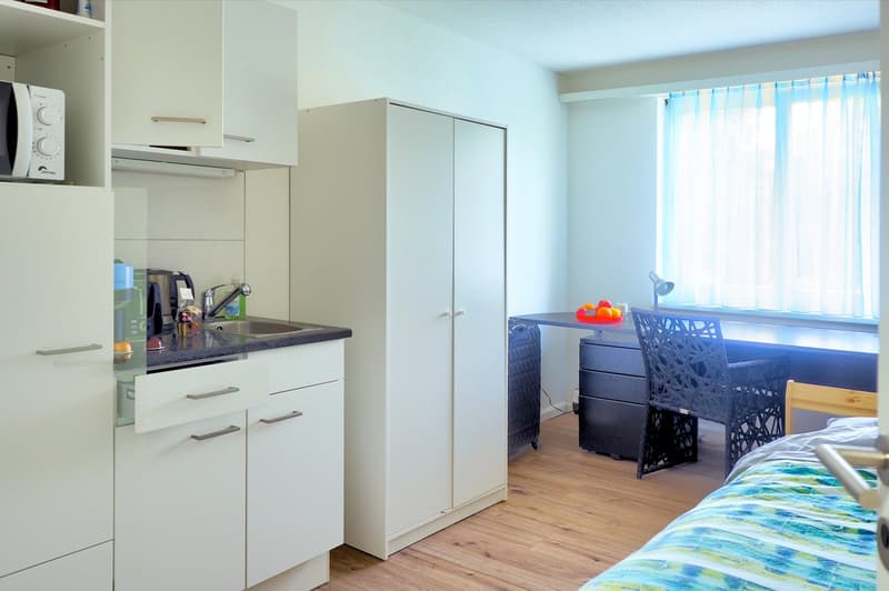 kitchen area with white cabinets, microwave, sink, drawers, desk, chair, bed, window