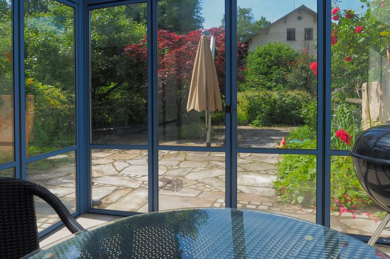 a room with large windows overlooking a stone paved area with plants and flowers, with a small outdoor table and a chair, and an outdoor umbrella