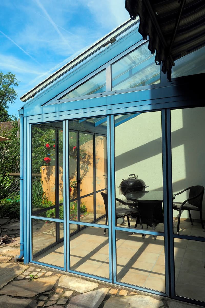 Enclosed patio space with glass doors and roof, tables and chairs, grill, stone floor, flowers, garden