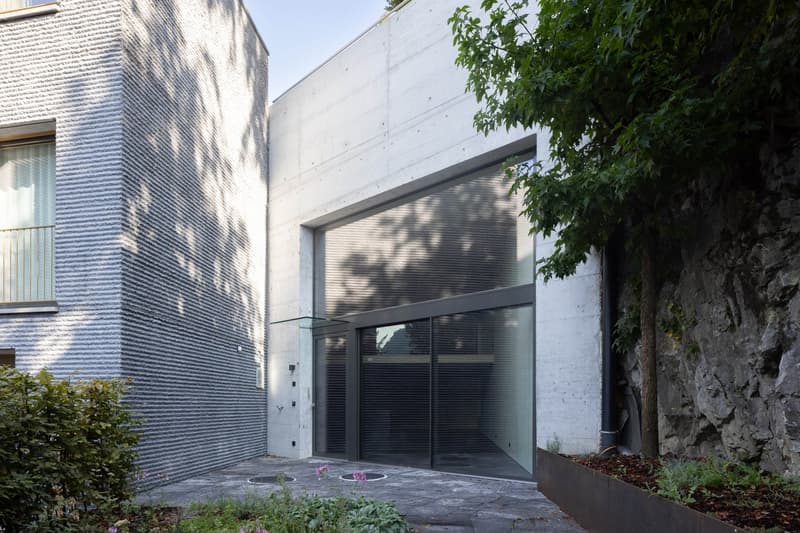 The image shows a modern, two-story building with a concrete exterior and large glass windows. There is a garage door visible on the ground floor, indicating the presence of a garage. The building is surrounded by trees and greenery, giving it a natural,