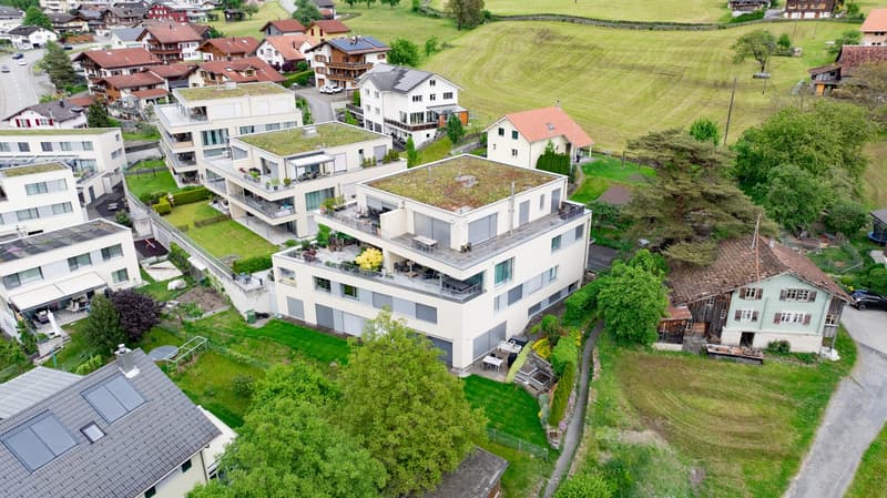 Multi-story apartment building with balconies, raised ground floor, and parking spaces. The building is surrounded by green spaces and other residential buildings in a scenic, rural setting.