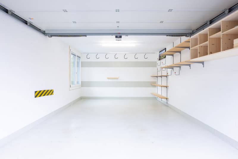 Spacious garage with white walls, overhead storage shelves, and a window. The garage has a concrete floor and appears to be well-lit.