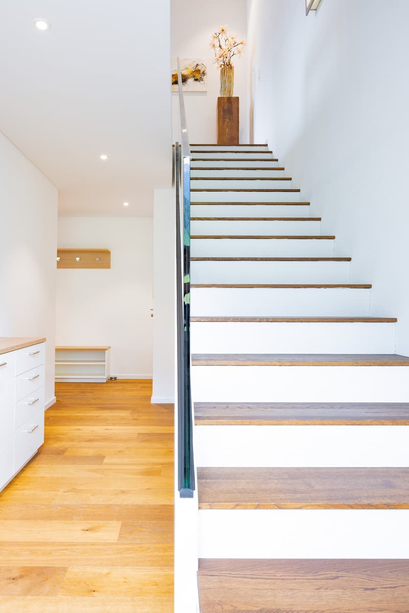 The image shows a modern and minimalist interior design with a wooden staircase leading to an upper level. The staircase features glass panels and wooden steps, creating a sleek and elegant look. There is a wooden vase with decorative branches placed on a