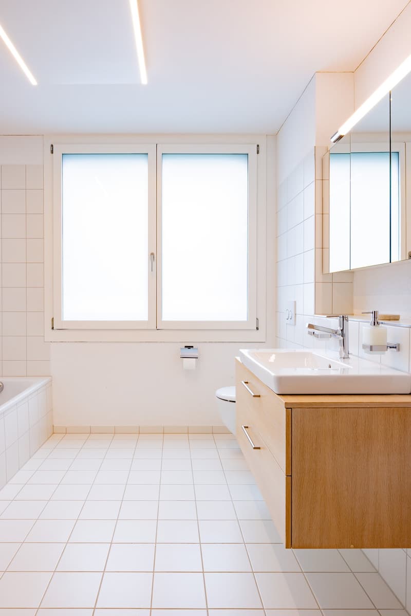 The image shows a modern and spacious bathroom with large windows, a white tiled floor, a wooden vanity unit with a vessel sink, and a mirror cabinet above the sink. The room appears to be well-lit and has a clean, minimalist design.