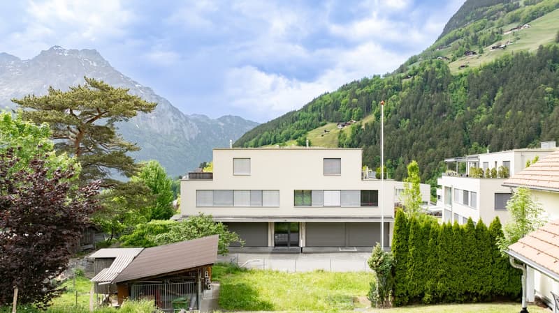 A modern, multi-story building with a minimalist design, surrounded by lush greenery and mountains in the background. The building has large windows and a balcony, suggesting it may be a residential or commercial property with a scenic view of the natural