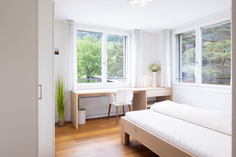 Spacious bedroom with large windows offering a scenic view of the surrounding greenery. The room features a wooden bed frame, a desk with chairs, and a potted plant, creating a bright and airy atmosphere.